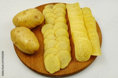 Raw potato tubers, raw potato slices and potato chips lie on a wooden board