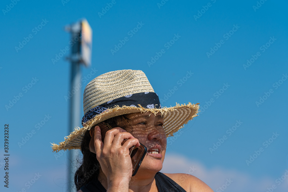 Closeup picture of a middle-aged Asian woman with a straw hat speaking in a smartphone a hot summer day. Bright blue sky in the background