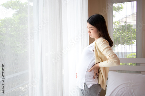 Young beautiful pregnant asian woman standing and touching belly and back near the window in bedroom. Pregnant women with low back pain, stress, anxiety concept.