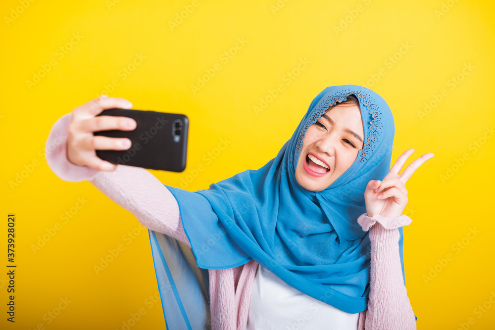 Asian Muslim Arab, Portrait of happy beautiful young woman Islam religious wear veil hijab funny smile she taking making selfie with smart mobile phone, studio shot isolated on yellow background Asian Muslim Arab, Portrait of happy beautiful young woman Islam religious wear veil hijab funny smile she taking making selfie with smart mobile phone, studio shot isolated on yellow background
