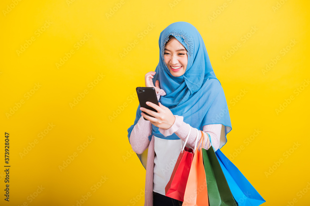 Asian Muslim Arab, Portrait of happy beautiful young woman Islam religious wear veil hijab funny smile she using smartphone on hand and hold shopping bags, studio shot isolated on yellow background Asian Muslim Arab, Portrait of happy beautiful young woman Islam religious wear veil hijab funny smile she using smartphone on hand and hold shopping bags, studio shot isolated on yellow background