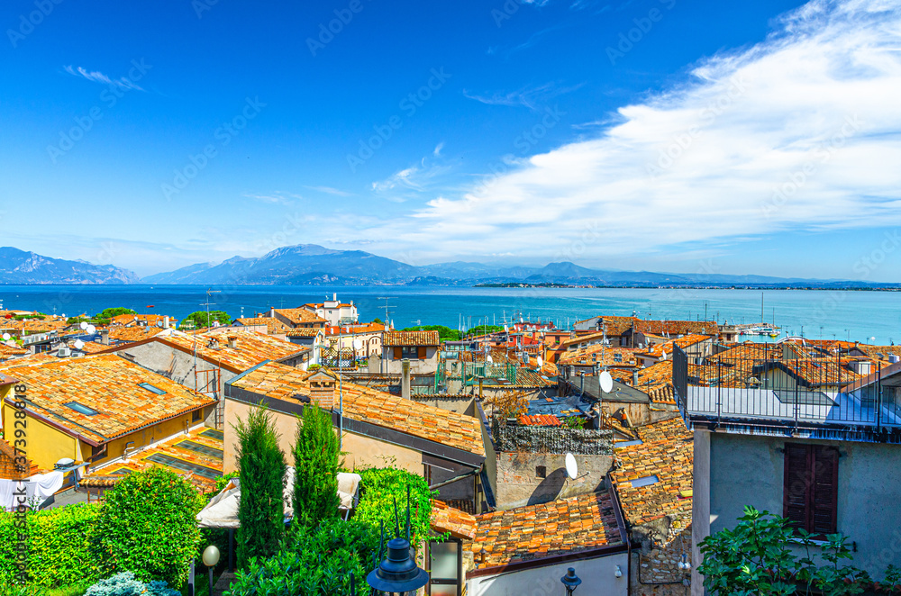 Aerial panoramic view of Desenzano del Garda town with red tiled roof buildings, Garda Lake water surface, Monte Baldo mountain range, Sirmione peninsula, blue sky background, Lombardy, Northern Italy