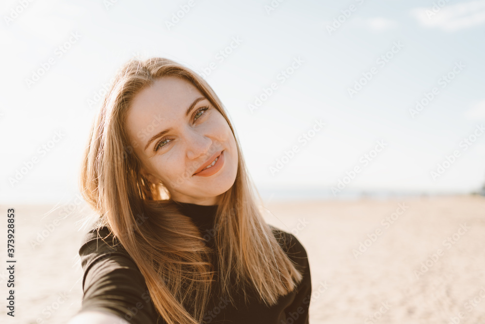 Pretty female with long hair takes selfie on smartphone on beach in summer or autumn. Beautiful woman looking at camera, tilt head and smiling in sunny day in ocean or sea coastline