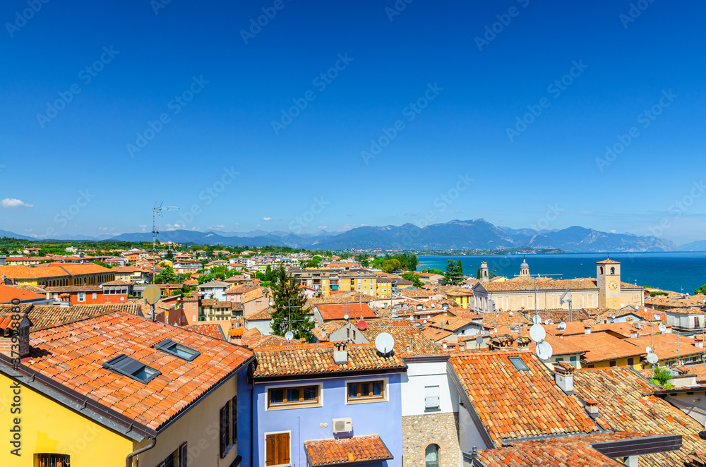 Aerial panoramic view of Desenzano del Garda town with bell tower of Duomo di Santa Maria Maddalena Cathedral church, red tiled roof buildings, Garda Lake, mountain range, Lombardy, Northern Italy