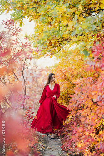 Red silk evening dress, long train fly, wind motion, walk autumn park. Pretty girl in a red long dress walks in the park.