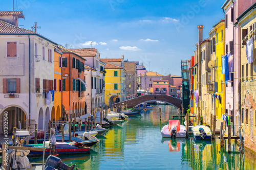 Chioggia cityscape with narrow water canal Vena with moored multicolored boats between old colorful buildings and brick bridge, blue sky background in summer day, Veneto Region, Northern Italy