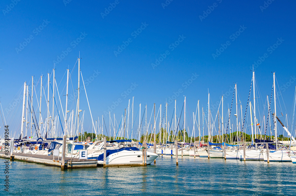 White yachts are moored on water of pier parking in marina port harbour of Sottomarina town in summer day, blue sky background, Veneto Region, Northern Italy