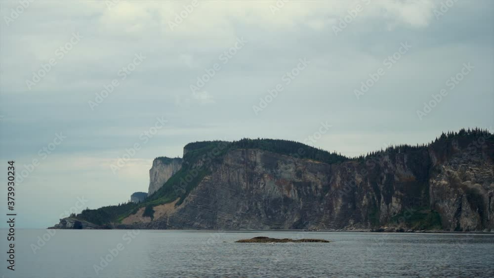 Serene Calm Sea Waters sit in front of an Epic Huge Flora Covered Ocean Cliffside on an Overcast Wintery Day at Forillon National Park, Canada. Serene Calm Sea Waters sit in front of an Epic Huge Flora Covered Ocean Cliffside on an Overcast Wintery Day at Forillon National Park, Canada.