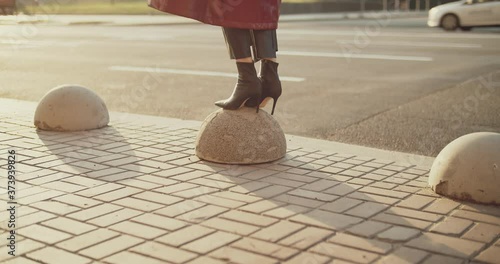 View of a woman legs wearing high heels jump on a sidewalk, making two steps. Balancing on a round surface near the road. Morning street full of cars
