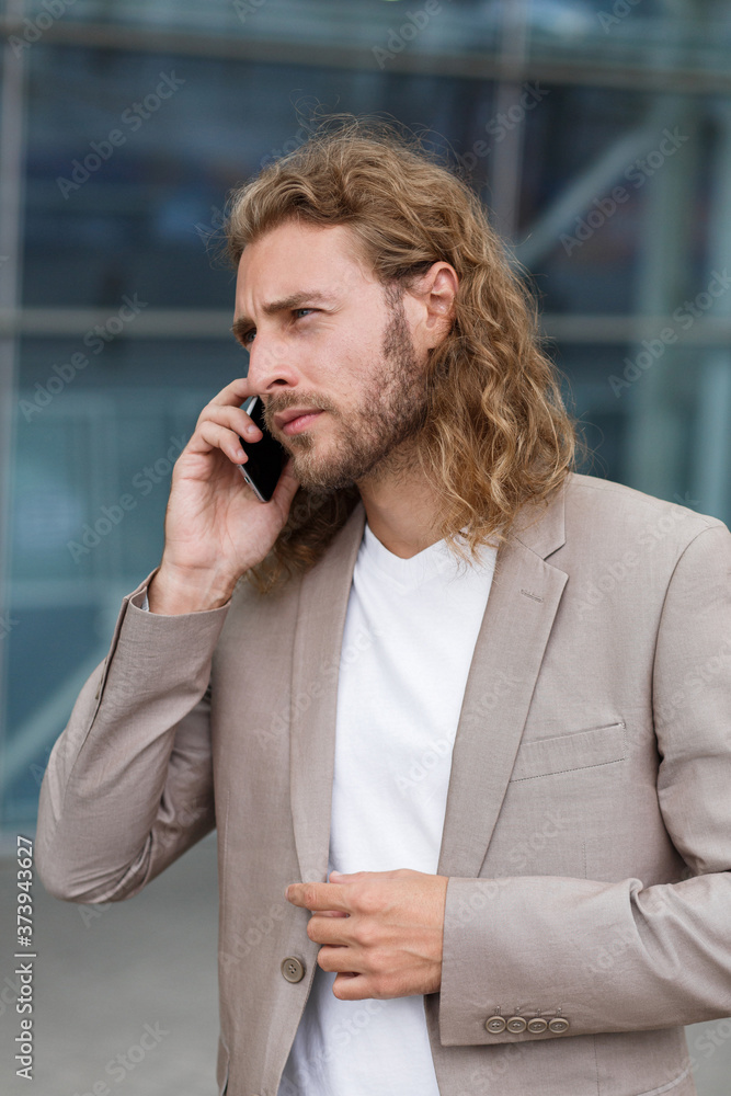 Young businessman talking on the phone, standing on city street near business center. Successful manager makes a call. Portrait of handsome curly smiling man in casual wear holding smartphone. Young businessman talking on the phone, standing on city street near business center. Successful manager makes a call. Portrait of handsome curly smiling man in casual wear holding smartphone.