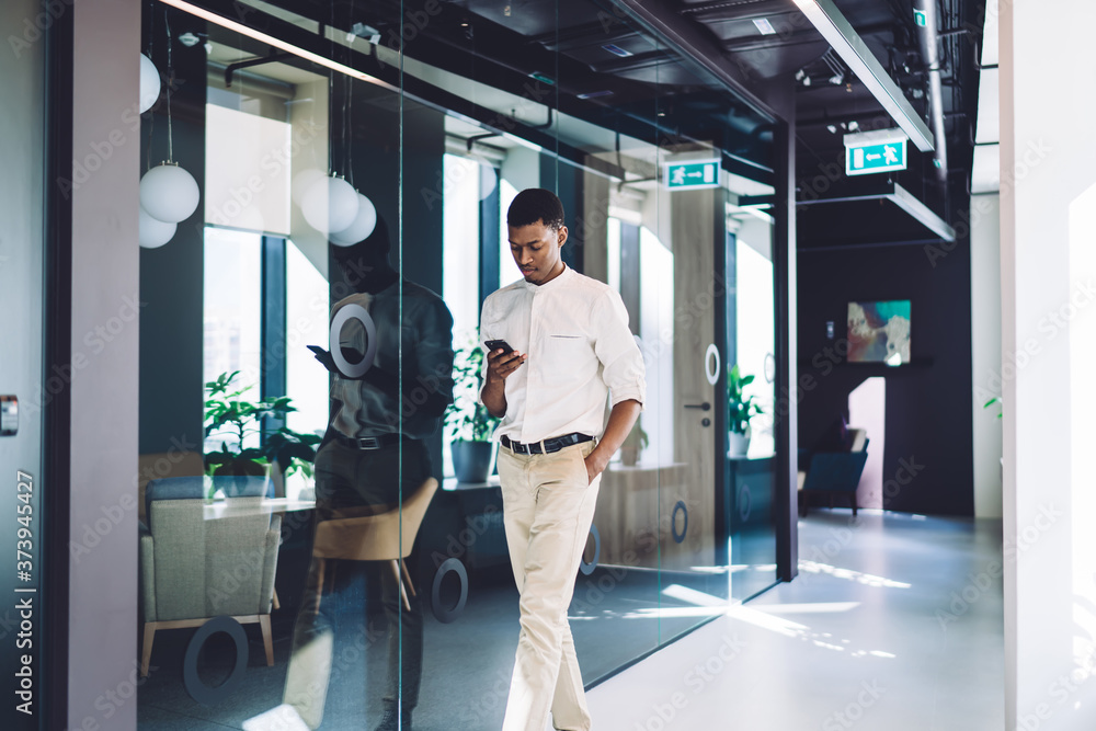 Fototapeta premium Focused ethnic businessman texting smartphone and walking along office hall