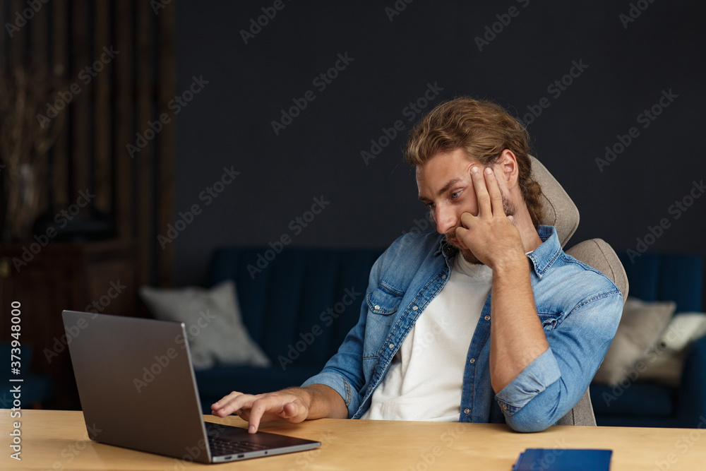 Young handsome curly smiling man with long hair working in home office with laptop. Business portrait of handsome manager sitting at workplace. Studying online, online courses. Business concept. Young handsome curly smiling man with long hair working in home office with laptop. Business portrait of handsome manager sitting at workplace. Studying online, online courses. Business concept.
