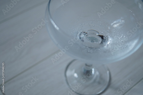 The cocktail glass stands on a light wooden plank surface. A glass in the right part of the frame, with sparkling water in it