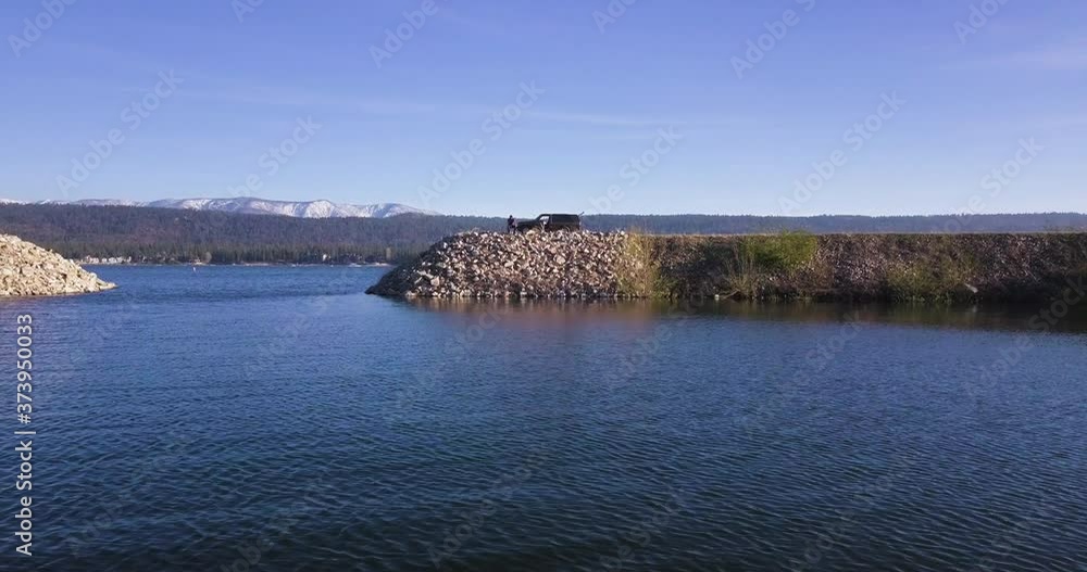 Fisherman sits on dyke with parked car behind him, drinking beer, waiting for fish to bite, sunny summer day, aerial shot forward Fisherman sits on dyke with parked car behind him, drinking beer, waiting for fish to bite, sunny summer day, aerial shot forward