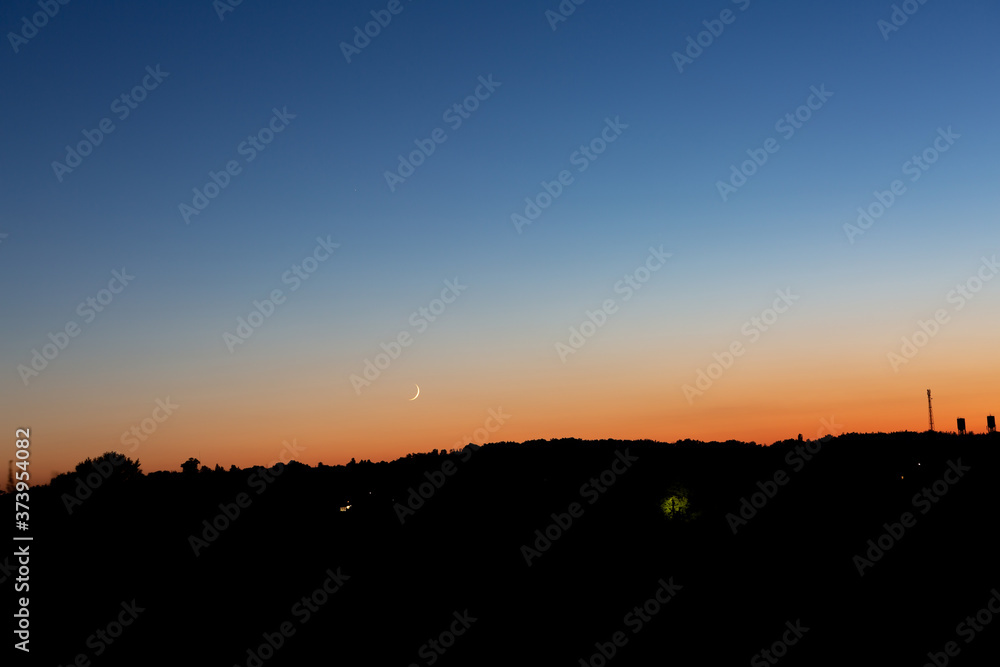 A young moon on an orange-blue sunset sky over a dark forest. Horizontal orientation. High quality photo