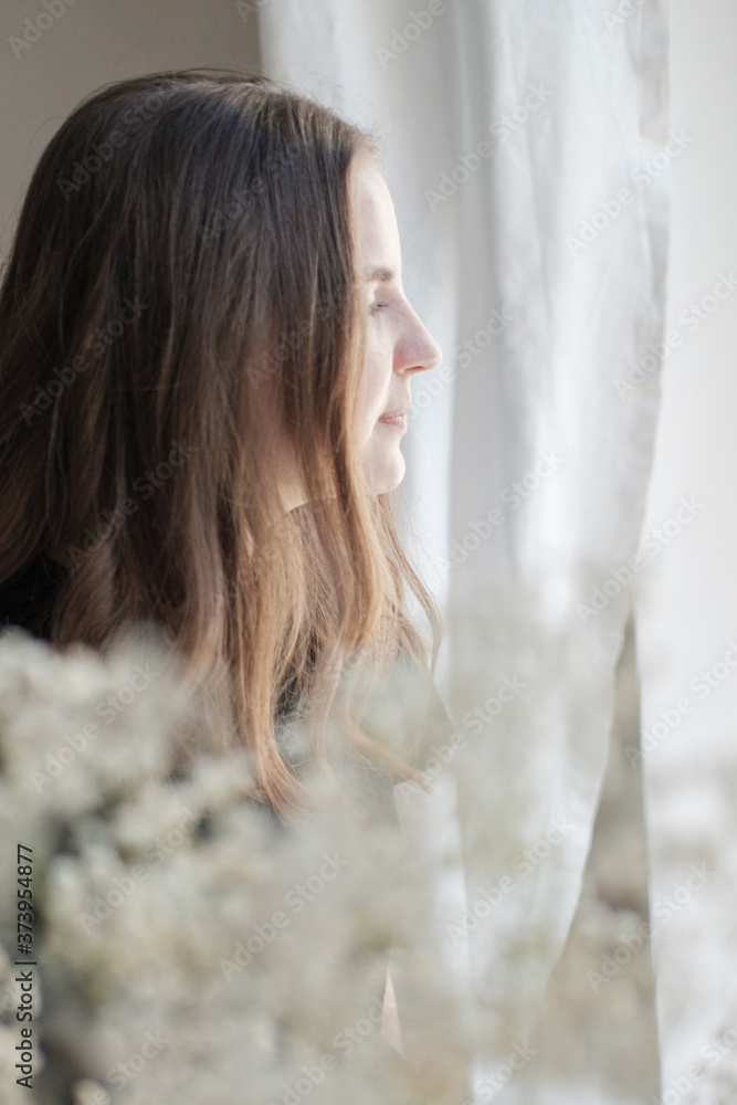 Portrait of young beautiful woman in black T-shirt by the window through white flowers. Sad, pensive girl is lost in her thoughts. Mindfulness concept. Portrait of young beautiful woman in black T-shirt by the window through white flowers. Sad, pensive girl is lost in her thoughts. Mindfulness concept.