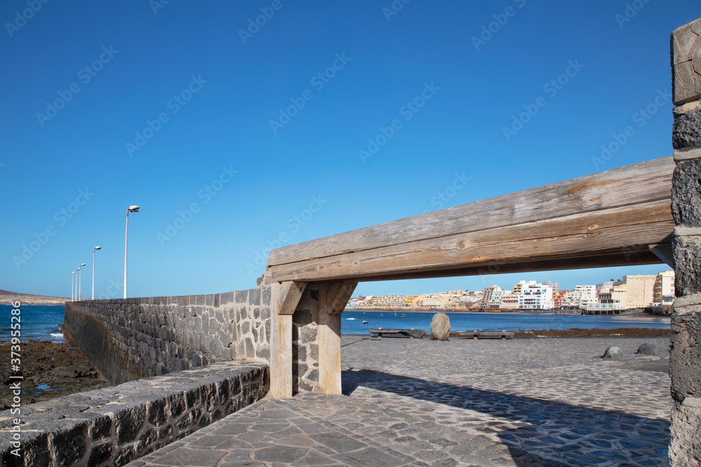 Fototapeta premium Views of El Medano town framed by a narrow wooden gangway and cobblestone pathways, towards the bay and residences behind, hotels and restaurants, beaches and coast, in Tenerife, Canary Islands, Spain