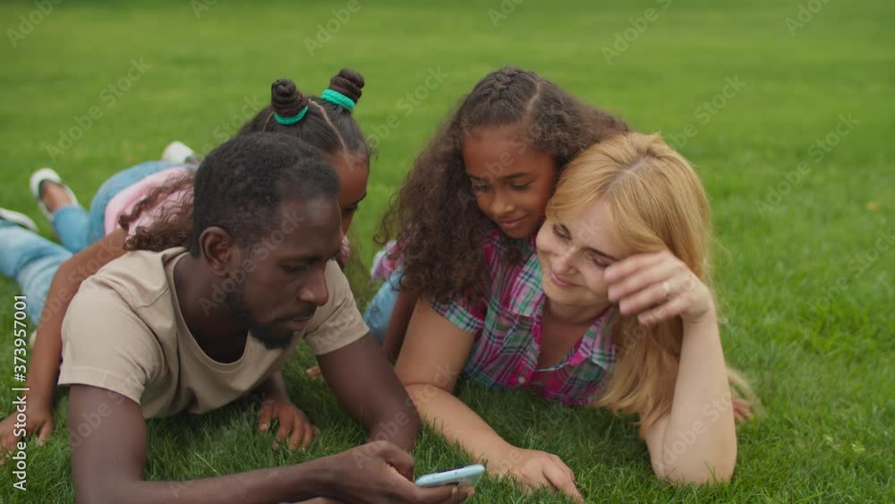 Joyful united multiethnic family with two adorable elementary age daughters lying on green grass, browsing social media content online on smart phone and talking while relaxing in summer nature.