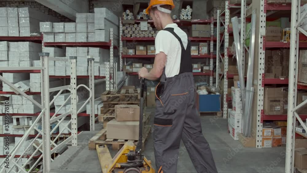 A factory worker in uniform is loading the cardboard boxes on a pallet truck. He is pushing the steering up and down. A factory worker in uniform is loading the cardboard boxes on a pallet truck. He is pushing the steering up and down.