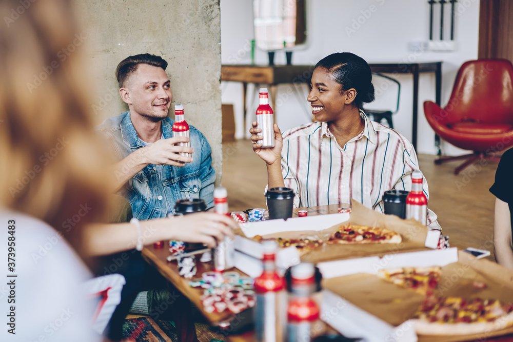 Fototapeta premium Hipster friends sitting at table with delivered pizza and alcohol drinks discussing board game with chips for playing poker during weekend, multicultural people enjoying gaming entertainment
