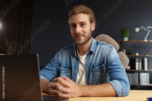 Young handsome curly smiling man with long hair working in home office with laptop. Business portrait of handsome manager sitting at workplace. Studying online, online courses. Business concept.