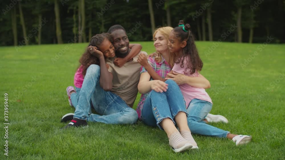 Two cute elementary age mixed race daughters running and embracing with love multiracial parents seated on green grass, expressing joy, happiness and togetherness while diverse family relaxing outdoor Two cute elementary age mixed race daughters running and embracing with love multiracial parents seated on green grass, expressing joy, happiness and togetherness while diverse family relaxing outdoor
