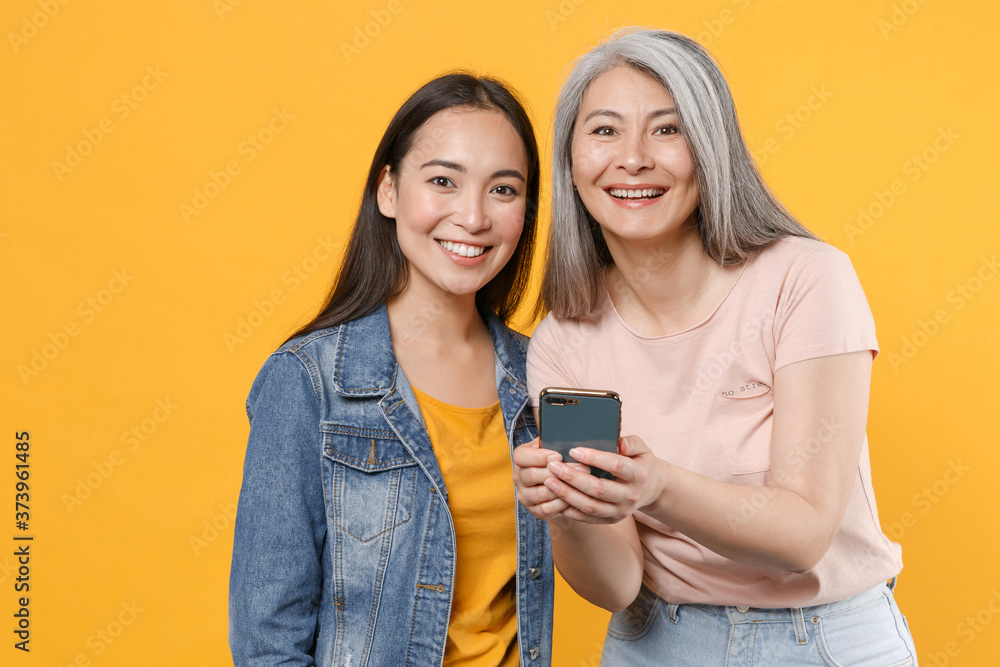 Smiling funny family asian female women gray-haired mother brunette daughter in casual clothes posing using mobile cell phone typing sms message isolated on yellow color background studio portrait. Smiling funny family asian female women gray-haired mother brunette daughter in casual clothes posing using mobile cell phone typing sms message isolated on yellow color background studio portrait.