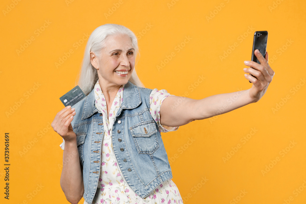 Smiling elderly gray-haired female woman wearing casual dress denim waistcoat posing doing selfie shot on mobile phone hold credit bank card isolated on yellow color wall background studio portrait. Smiling elderly gray-haired female woman wearing casual dress denim waistcoat posing doing selfie shot on mobile phone hold credit bank card isolated on yellow color wall background studio portrait.