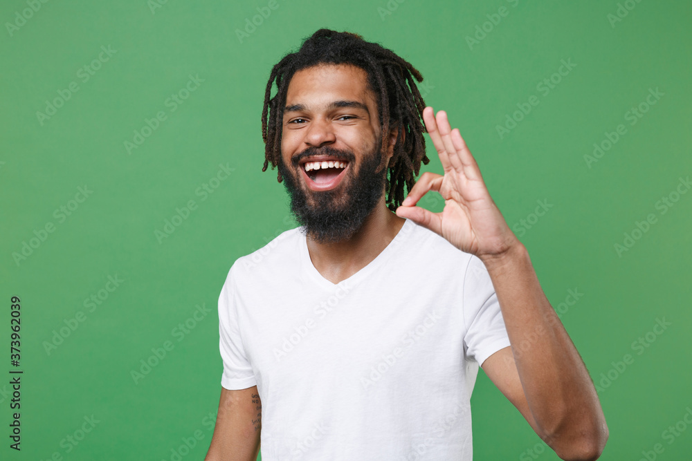 Cheerful funny young african american man guy wearing white casual t-shirt posing isolated on green wall background studio portrait. People sincere emotions lifestyle concept. Showing OK gesture. Cheerful funny young african american man guy wearing white casual t-shirt posing isolated on green wall background studio portrait. People sincere emotions lifestyle concept. Showing OK gesture.