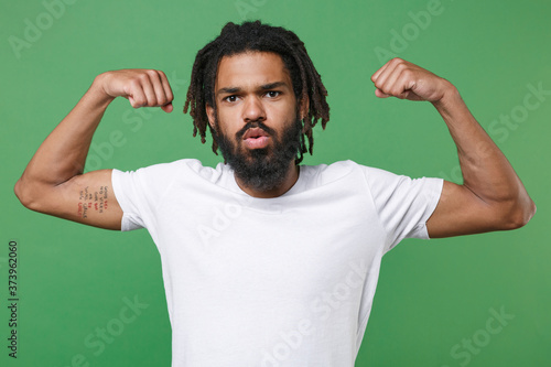 Strong amazed young african american man guy 20s wearing white casual t-shirt posing showing biceps muscles spreading hands looking camera isolated on green color wall background studio portrait.