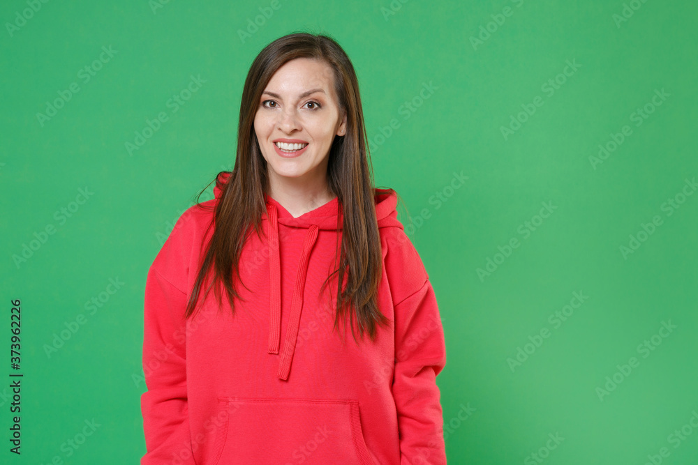 Smiling cheerful beautiful attractive young brunette woman 20s wearing bright red casual streetwear hoodie posing standing and looking camera isolated on green color wall background studio portrait. Smiling cheerful beautiful attractive young brunette woman 20s wearing bright red casual streetwear hoodie posing standing and looking camera isolated on green color wall background studio portrait.
