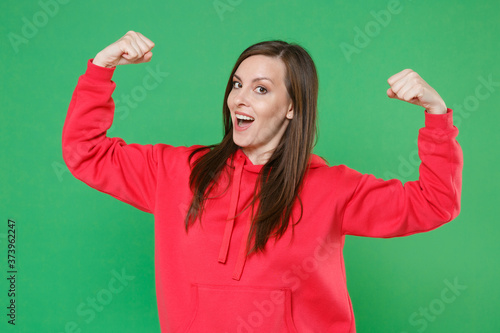Strong cheerful funny young brunette woman 20s wearing bright red casual streetwear hoodie posing showing biceps muscles looking camera isolated on green color wall background studio portrait.