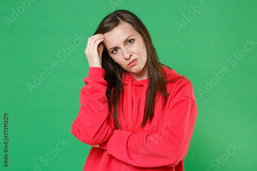Confused displeased puzzled young brunette woman 20s wearing bright red casual streetwear hoodie posing put hand on head looking camera isolated on green color wall background studio portrait.