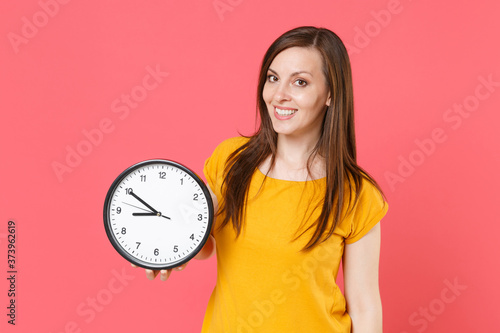 Smiling beautiful attractive charming young brunette woman 20s in yellow casual t-shirt posing standing holding in hands clock looking camera isolated on pink color wall background studio portrait.