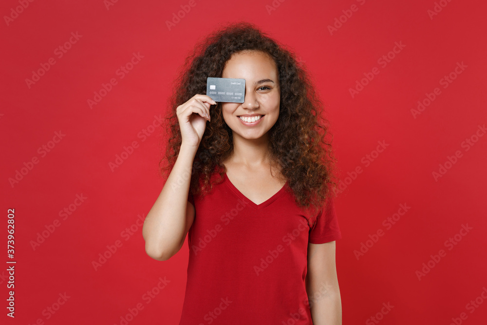 Obraz premium Smiling young african american woman girl in casual t-shirt posing isolated on red wall background studio portrait. People lifestyle concept. Mock up copy space. Covering eye with credit bank card.