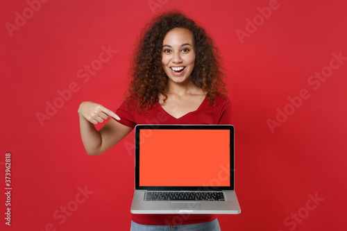 Funny young african american woman girl in casual t-shirt isolated on red background. People lifestyle concept. Mock up copy space. Pointing index finger on laptop pc computer with blank empty screen. © ViDi Studio