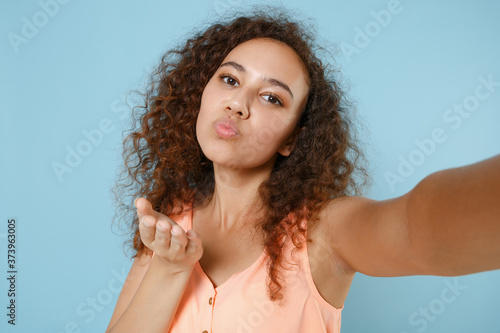 Close up of pretty african american girl in pastel summer clothes isolated on blue background. People lifestyle concept. Mock up copy space. Doing selfie shot on mobile phone, blowing send air kiss.