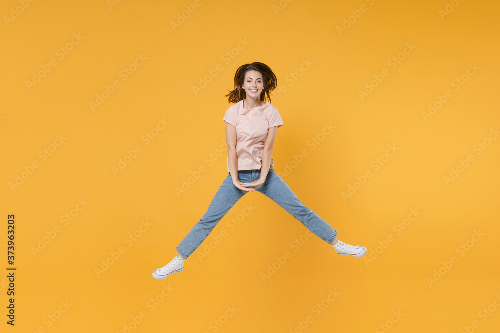 Full length portrait of smiling cheerful funny young woman 20s wearing pastel pink casual t-shirt posing jumping spreading legs looking camera isolated on bright yellow color wall background studio. Full length portrait of smiling cheerful funny young woman 20s wearing pastel pink casual t-shirt posing jumping spreading legs looking camera isolated on bright yellow color wall background studio.