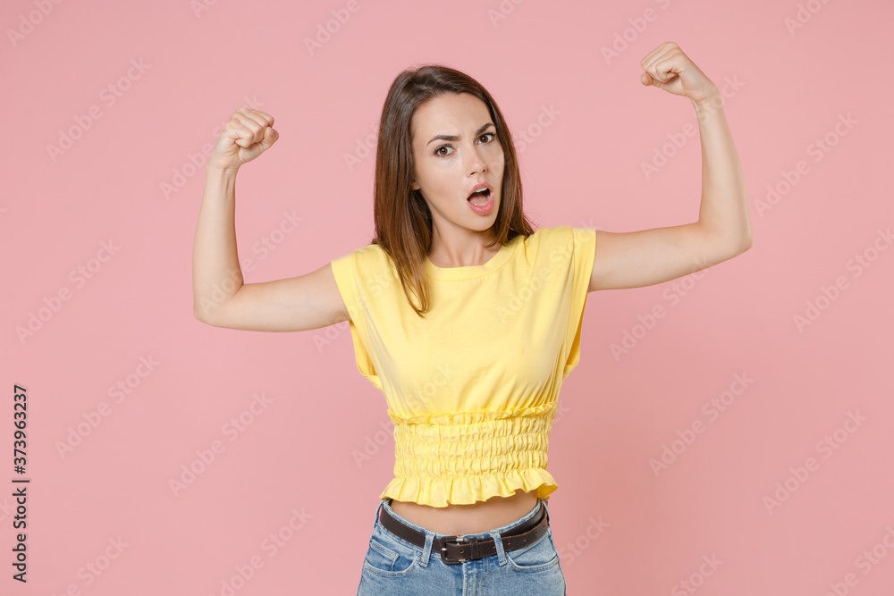Shocked strong young brunette woman 20s wearing yellow casual t-shirt posing standing showing biceps muscles spreading hands looking camera isolated on pastel pink color background studio portrait. Shocked strong young brunette woman 20s wearing yellow casual t-shirt posing standing showing biceps muscles spreading hands looking camera isolated on pastel pink color background studio portrait.