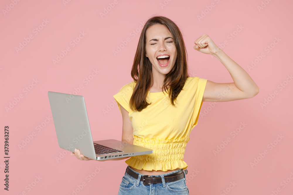 Happy young brunette woman wearing yellow casual t-shirt posing stand working on laptop pc computer doing winner gesture keeping eyes closed isolated on pastel pink color background studio portrait. Happy young brunette woman wearing yellow casual t-shirt posing stand working on laptop pc computer doing winner gesture keeping eyes closed isolated on pastel pink color background studio portrait.
