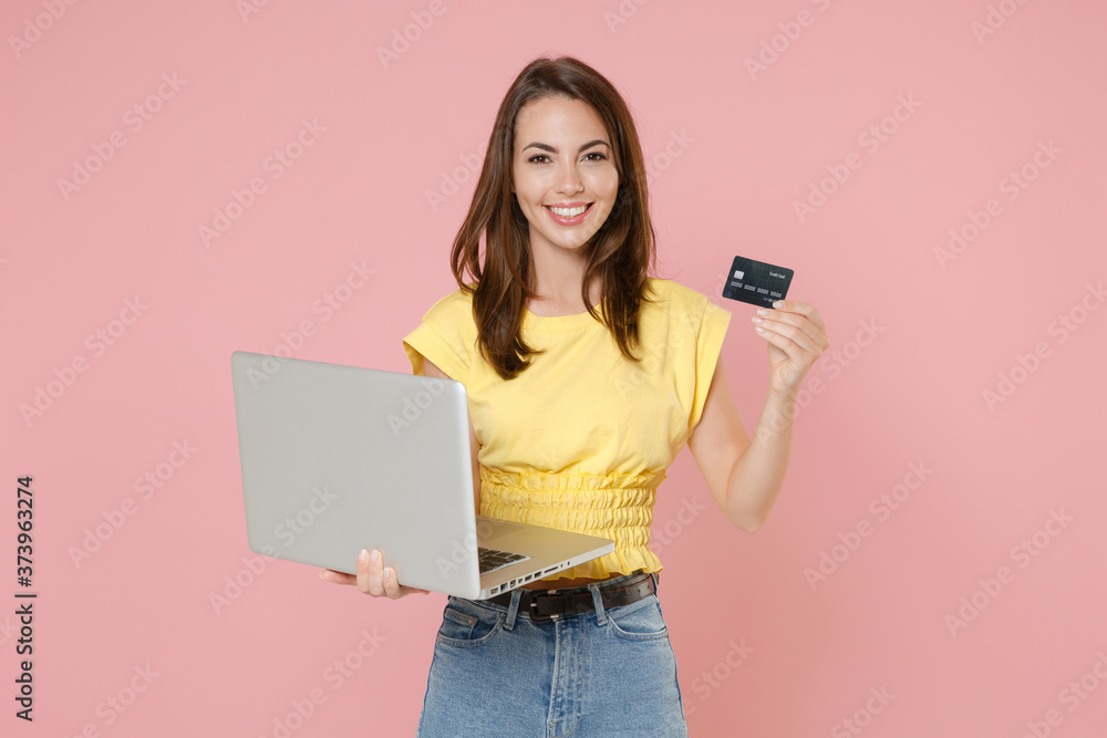 Naklejka premium Smiling young brunette woman wearing yellow casual t-shirt posing standing working on laptop pc computer hold credit bank card looking camera isolated on pastel pink color background studio portrait.