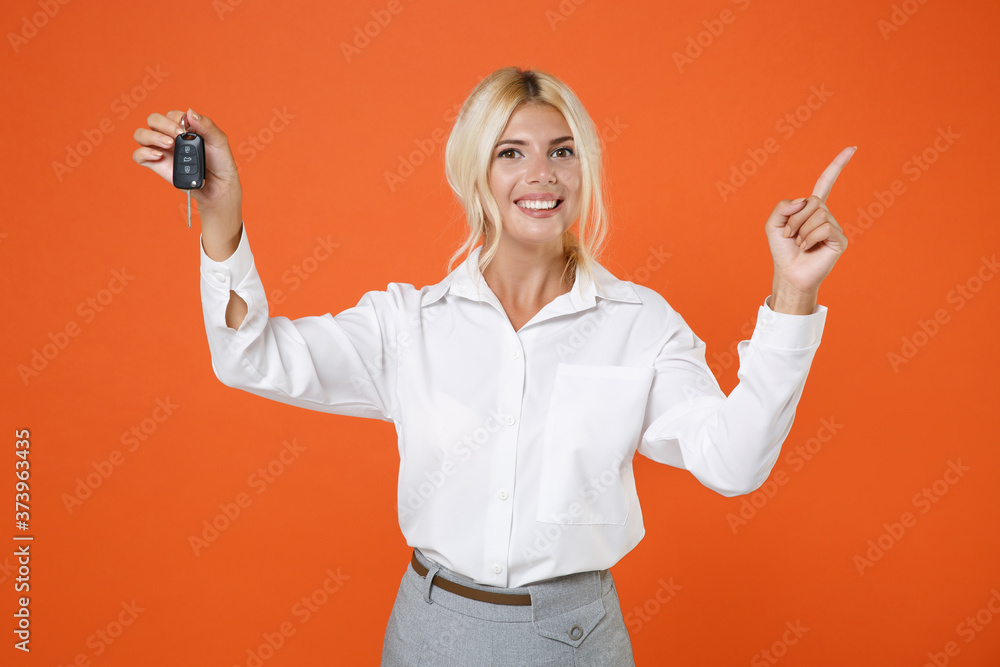Obraz premium Smiling beautiful young blonde female business woman in white shirt hold car keys pointing index finger up on mock up copy space looking camera isolated on orange color background studio portrait.