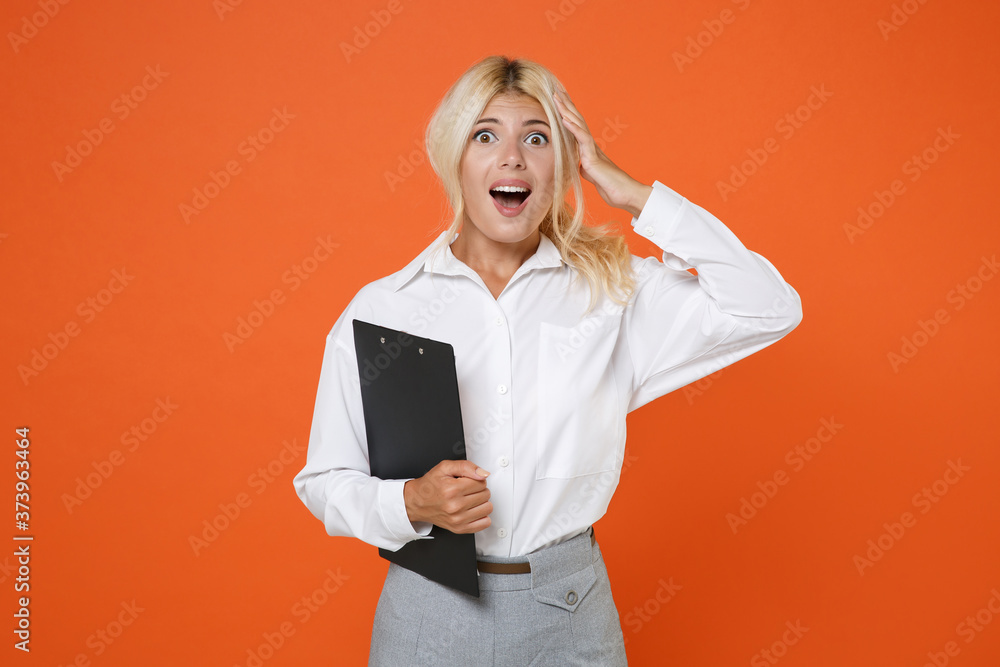 Shocked young blonde female business woman girl wearing white shirt posing hold clipboard with papers document put hand on head keeping mouth open isolated on orange color background studio portrait. Shocked young blonde female business woman girl wearing white shirt posing hold clipboard with papers document put hand on head keeping mouth open isolated on orange color background studio portrait.