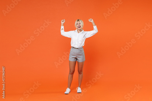 Full length portrait of happy joyful young blonde female business woman girl 20s in white shirt posing clenching fists doing winner gesture isolated on bright orange color wall background studio. © ViDi Studio