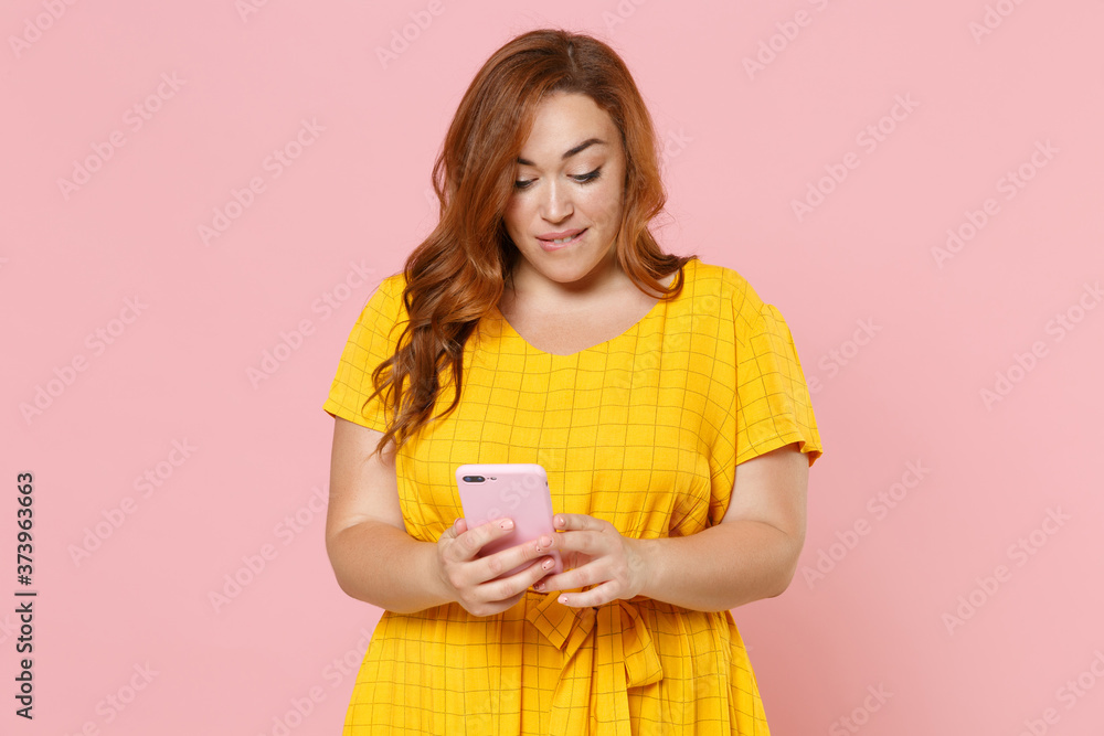 Pensive young redhead plus size body positive female woman 20s in yellow dress posing using mobile cell phone typing sms message biting lips isolated on pastel pink color background studio portrait. Pensive young redhead plus size body positive female woman 20s in yellow dress posing using mobile cell phone typing sms message biting lips isolated on pastel pink color background studio portrait.