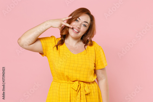 Smiling cheerful young redhead plus size body positive female woman girl 20s in yellow casual dress posing showing victory sign looking camera isolated on pastel pink color background studio portrait.