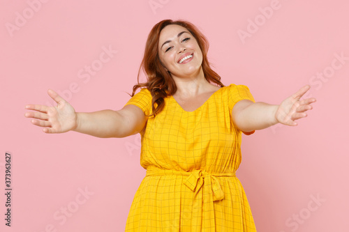 Smiling pretty young redhead plus size body positive female woman girl 20s in yellow dress posing reach out stretching hands looking camera isolated on pastel pink color background studio portrait.