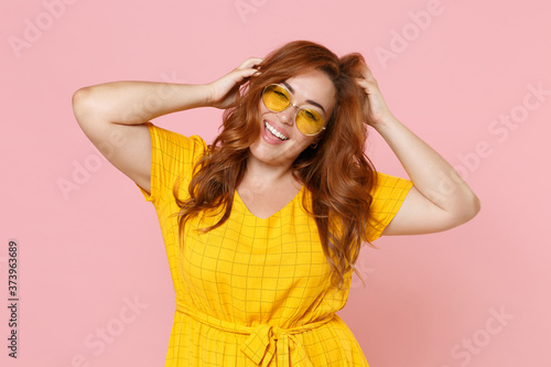 Cheerful funny beautiful young redhead plus size body positive female woman girl 20s in yellow dress eyeglasses posing put hands on head isolated on pastel pink color wall background studio portrait.