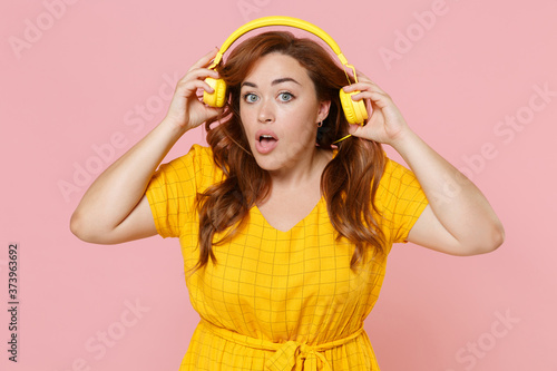 Shocked young redhead plus size body positive female woman 20s in yellow dress posing listening music with headphones keeping mouth open isolated on pastel pink color wall background studio portrait.