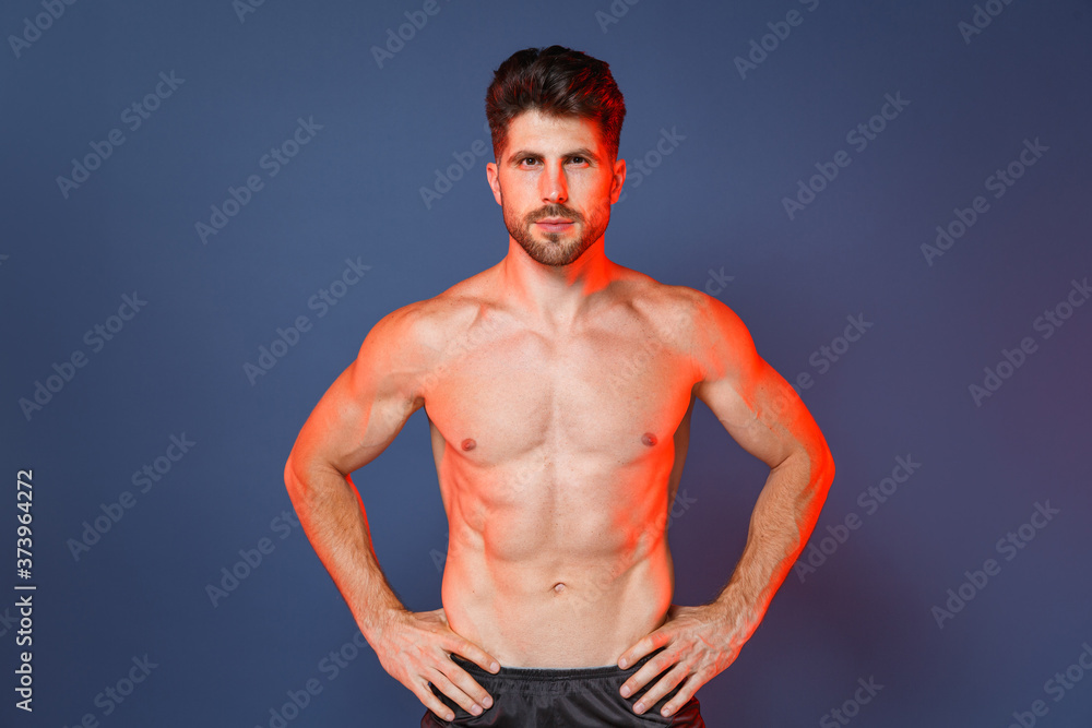 Fototapeta premium Handsome young bearded fitness sporty strong guy bare-chested muscular sportsman isolated on blue background studio. Workout sport motivation lifestyle concept. Standing with arms akimbo on waist.